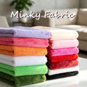Könnte beinhalten: Ein Stapel gefalteter Minky-Stoff in verschiedenen Farben, darunter Lavendel, Pink, Orange, Weiß, Grün, Oliv, Pfirsich, Hellrosa, Magenta, Rot, Dunkelrot und Schwarz. Der Stoff hat eine weiche, plüschige und flauschige Textur mit sichtbarem Flor.