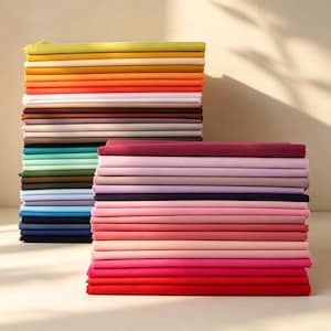Puede incluir: Dos pilas altas y ordenadas de cuadrados de tela doblados en un amplio espectro de colores. La pila de la izquierda presenta tonos amarillos, naranjas, blancos, marrones y azules, mientras que la derecha muestra tonos de rosa, magenta, morado y rojo.