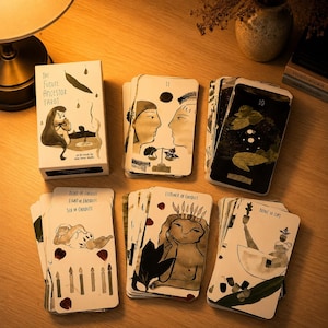 Könnte beinhalten: Ein Kartendeck The Future Ancestor Tarot mit einer weißen Schachtel, die eine grüne Blattillustration zeigt. Die Karten zeigen schwarz-weiße Tuschezeichnungen von Figuren, Kerzen und Pflanzen mit Texten wie Six of Candles und Nine of Cups.
