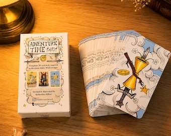 Op de afbeelding: Een 78-kaarten Adventure Time Tarot-deck met een witte doos met de titel, illustraties van Kracht, Gerechtigheid en Page van Kelken, en de naam van de kunstenaar. De waaier van kaarten toont een gouden kelk met een zwaard, bloemen en wolken.