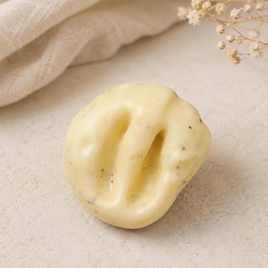 May include: A round, pale yellow bar of soap with a smooth, glossy surface and visible dark specks embedded throughout. The soap features a distinct, deep, curved indentation running across its center, creating a unique, organic, and rounded shape.
