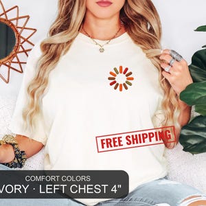 May include: An ivory short-sleeved t-shirt featuring a small, circular graphic on the left chest composed of twelve pill-shaped icons in shades of orange, brown, and green arranged in a loading-style pattern. The shirt has a crew neckline.