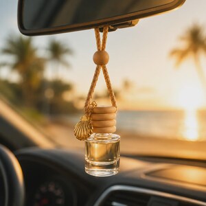 May include: A clear glass car air freshener bottle with a light wood cap, suspended by a braided tan cord. A gold-toned metal seashell charm hangs from the cord. The bottle contains a pale liquid and is hanging from a vehicle rearview mirror.