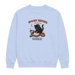 Puede incluir: Una sudadera azul claro de cuello redondo con un gráfico de un gato negro amasando masa sobre una tabla de madera. El texto dice SPOOKY BISCUITS bakery y WE KNEAD EM YOU NEED EM, junto a un cuenco pequeño, una calabaza y una luna creciente.