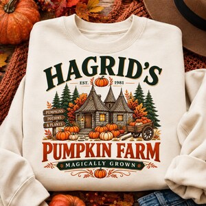 Puede incluir: Una sudadera de cuello redondo color crema con un estampado gráfico de una cabaña de piedra con techo cónico, rodeada de pinos y calabazas. El texto dice HAGRID'S EST. 1981 PUMPKIN FARM MAGICALLY GROWN, con un letrero de madera que enumera PUMPKINS, POTIONS & PLANTS.