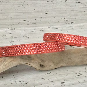 Puede incluir: Dos collares de cuero de color coral con filas de brillantes de imitaci&oacute;n. Los collares est&aacute;n sobre una superficie de madera clara.