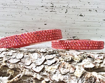 May include: Two pink rhinestone dog collars on a wooden surface. The collars are made of leather and have a row of pink rhinestones around the edge.