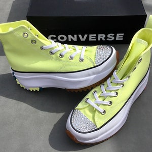 May include: A pair of yellow high-top Converse sneakers with a platform sole and a white rubber toe cap. The sneakers have white laces and a row of clear rhinestones on the toe cap. The sneakers are sitting on a gray surface. The word "CONVERSE" is visible on a black box in the background.