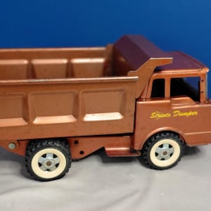 May include: A copper-colored metal toy dump truck with a large open cargo bed. The side of the truck features the yellow text "Stratos Dumper". It has four black wheels with white rims and a cabin with an open window design on the front side.