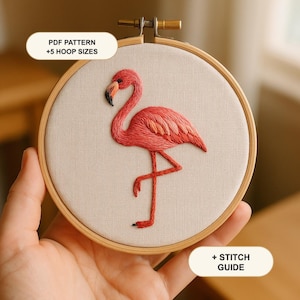 May include: A round wooden embroidery hoop containing off-white fabric with a hand-stitched pink flamingo design. Text on the image reads: PDF PATTERN +5 HOOP SIZES and + STITCH GUIDE. The flamingo features shades of pink and a black beak.