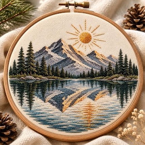 Op de afbeelding: Een ronde houten borduurring met een stoffen paneel voorzien van een gedetailleerd borduurwerk van een landschap. De scène toont een bergketen, een dennenbos en een zon, allemaal weerspiegeld in een kalm meer met blauwe en gouden draad.