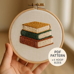 May include: A circular wooden embroidery hoop containing a fabric panel with a hand-stitched design of three stacked books in mustard yellow, dark green, and rust orange. A white circular overlay includes the text PDF PATTERN +4 HOOP SIZES.