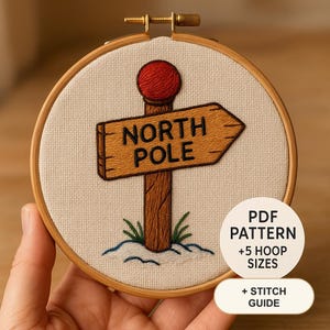 Könnte beinhalten: Ein Stickrahmen mit einem Stoffstück, das ein gesticktes braunes Holzschild zeigt, das nach rechts weist und mit NORTH POLE in schwarzem Garn beschriftet ist. Der Pfosten hat eine rote Kugel an der Spitze und steht in weißem Schnee mit grünem Gras. Text: PDF PATTERN +5 HOOP SIZES + STITCH GUIDE.