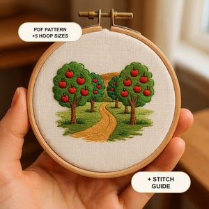 Puede incluir: Un bastidor de bordado con tela color crema que muestra un paisaje bordado de manzanos y un camino sinuoso. El texto "PDF PATTERN +5 HOOP SIZES" y "+ STITCH GUIDE" es visible. Los árboles tienen hojas verdes y manzanas rojas.