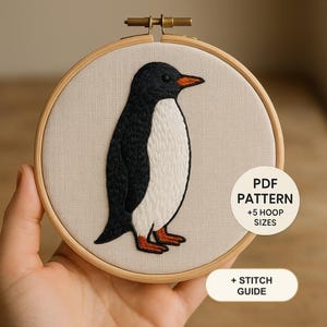Op de afbeelding: Een ronde houten borduurring met een beige stoffen paneel met een handgeborduurd pinguïnontwerp. De pinguïn is gemaakt met zwarte, witte en oranje draad. Een witte cirkelvormige overlay bevat de tekst PDF PATTERN +5 HOOP SIZES en + STITCH GUIDE.