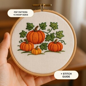 Puede incluir: Un bastidor de bordado redondo de madera que contiene tela blanca con un diseño bordado a mano de tres calabazas naranjas y hojas verdes. El texto dice PDF PATTERN +5 HOOP SIZES y + STITCH GUIDE. El diseño presenta un trabajo de hilo detallado.
