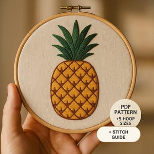 Op de afbeelding: Een ronde houten borduurring met een stoffen paneel met een handgeborduurd ananasontwerp in geel en donkergroen. Een rond wit label bevat de tekst PDF PATTERN +5 HOOP SIZES en + STITCH GUIDE in zwarte letters.