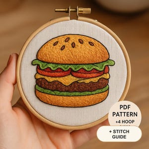 May include: A round wooden embroidery hoop containing a fabric panel with a hand-stitched hamburger design featuring a golden bun, green lettuce, red tomato, yellow cheese, and a brown patty. A circular label reads PDF PATTERN +4 HOOP SIZES + STITCH GUIDE.