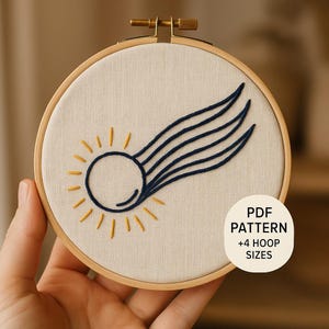 May include: A circular wooden embroidery hoop containing off-white fabric with a stitched design of a sun with a dark blue comet tail. The design features yellow rays and dark blue lines. A white circular label reads PDF PATTERN +4 HOOP SIZES.