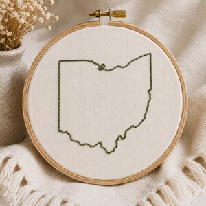 Pode incluir: Um bastidor de bordado circular de madeira contendo tecido branco sujo. O tecido apresenta o contorno do estado de Ohio bordado com linha verde escura em ponto atrás. O bastidor possui um parafuso de aperto de metal dourado no topo.