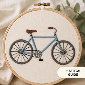 Op de afbeelding: Een ronde houten borduurring met een stoffen paneel waarop een handgeborduurd blauw fietsontwerp staat. De fiets heeft een bruin zadel, grijze banden met zwarte spaken en een pedaal. Een wit label bevat de tekst + STITCH GUIDE.
