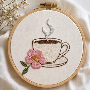 Op de afbeelding: Een ronde houten borduurring met een wit stoffen paneel waarop een handgeborduurd ontwerp van een crèmekleurige koffiekop op een schoteltje staat. De kop bevat donkerbruine koffie met lichtgrijze stoom en een roze bloem met groene blaadjes.