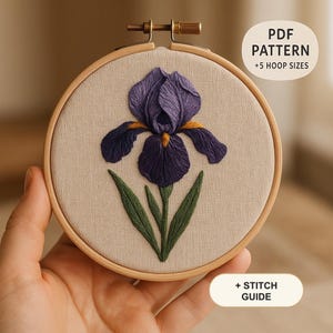 Könnte beinhalten: Ein runder Stickrahmen aus Holz mit einem beigen Stoffeinsatz, der eine handgestickte violette Irisblüte mit grünen Blättern und gelben Akzenten zeigt. Der Text auf dem Bild lautet PDF PATTERN +5 HOOP SIZES und + STITCH GUIDE.