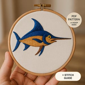 May include: A round wooden embroidery hoop containing white fabric with a hand-stitched blue and gold marlin fish design. The text on the image reads PDF PATTERN +4 HOOP SIZES and + STITCH GUIDE. The fish features a prominent dorsal fin and bill.