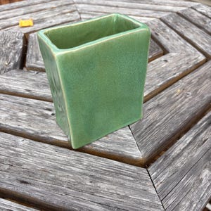 May include: A rectangular ceramic vase with a glossy, light green glaze. The vase features a smooth, uniform surface with slightly rounded corners and a wide, open top, showcasing a consistent finish throughout the entire exterior structure.