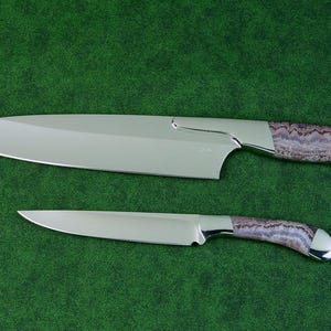 Peut inclure: Deux couteaux de cuisine avec des lames en métal argenté poli et des manches présentant un motif texturé semblable à de la pierre dans des tons de violet, blanc et gris. Le plus grand couteau a une lame large, tandis que le plus petit est plus fin.