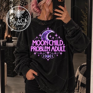 Könnte beinhalten: Ein schwarzes Langarm-Sweatshirt mit einer abgenutzten, verwaschenen Textur. Auf der Vorderseite befindet sich ein Grafikdesign mit einem violetten Halbmond, Sternen und dem Text "MOON CHILD, PROBLEM ADULT." in leuchtendem Neon-Pink.