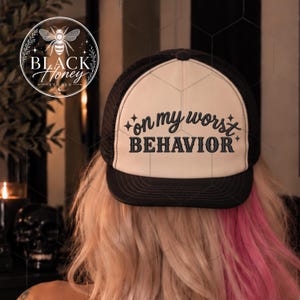 May include: A black and white trucker hat featuring a foam front panel with the embroidered text "on my worst BEHAVIOR" in a dark font, accented by small star symbols. The hat has a black mesh back, a black brim, and a black button on top.