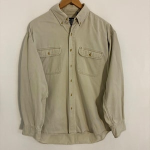 Puede incluir: Una camisa de manga larga con botones en color beige sólido. La camisa presenta un cuello abotonado, una tapeta central con botones y dos bolsillos en el pecho con solapas abotonadas. La tela tiene una textura visible y la marca Gap es visible.
