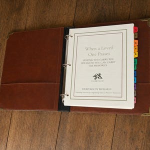 May include: A brown three-ring binder with metal corner protectors containing white pages. The cover page reads: When a Loved One Passes, Helping You Carry the Details So You Can Carry the Memories, Heritage by Morales. Includes colorful index tabs.