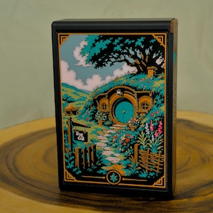 May include: A rectangular black box featuring a colorful illustration of a round green door set into a grassy hill with a large tree above. A sign near the path reads Bag End. The artwork uses shades of teal, green, and orange with gold accents.