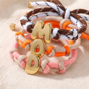 May include: A stack of three braided cord bracelets featuring gold-toned metal letter charms that spell out the word MOD. The bracelets are crafted from white and brown, orange and beige, and pink and beige cords with small gold-toned metal end caps.