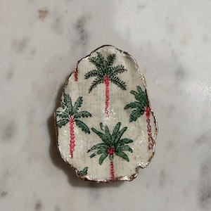 Puede incluir: Un pequeño plato decorativo con forma de concha irregular y superficie blanca texturizada que presenta un patrón repetitivo de palmeras verdes con troncos rosas. Los bordes del plato están acabados con un borde metálico dorado decorativo.