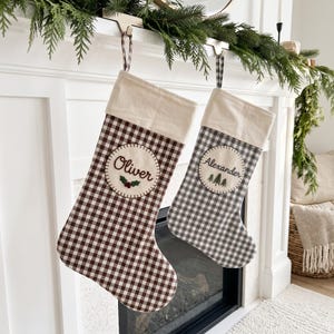 Peut inclure: Deux chaussettes de Noël à motif vichy, l'une marron et l'autre grise, dotées d'un revers crème et d'un écusson circulaire. La chaussette marron porte le nom Oliver et du houx brodés, tandis que la grise affiche le nom Alexander et des sapins.