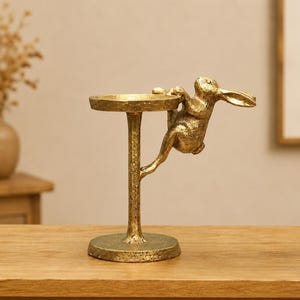 Puede incluir: Un soporte de pedestal de metal dorado con acabado texturizado y una bandeja circular superior. Una figura de conejo esculpida está unida al lado del poste central, pareciendo trepar hacia la bandeja con sus patas delanteras tocando el borde.