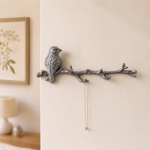 May include: A silver-toned metal wall hook shaped like a tree branch with a bird perched on the left side. The branch features multiple small hooks for hanging items, and a thin gold-coloured chain necklace is currently hanging from one hook.