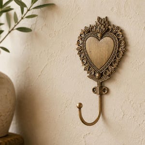 Peut inclure: Un crochet mural doté d'une plaque décorative en métal couleur laiton en forme de cœur, avec une bordure en filigrane ornée et un motif de flamme au sommet. Le crochet s'étend depuis la base du cœur, qui présente un petit détail ailé.