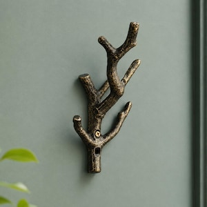 Puede incluir: Un gancho de pared con forma de rama de árbol con un acabado texturizado de color bronce oscuro. El gancho presenta múltiples puntas curvadas hacia arriba para colgar objetos y está fijado a la pared con un tornillo metálico visible.