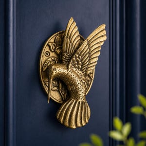 Op de afbeelding: Een messingkleurige metalen deurklopper in de vorm van een vliegende kolibrie. De vogel is afgebeeld met de vleugels omhoog gespreid en een lange, puntige snavel, gemonteerd op een ronde achterplaat met een reliëf van bladeren en bloemen.