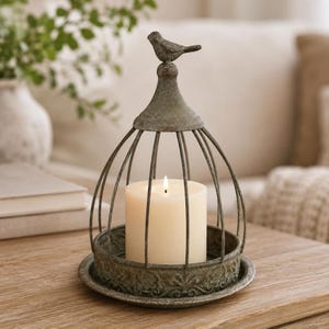 May include: A dome-shaped metal candle holder with a weathered green finish, featuring a small bird figurine perched on top. The cage-like structure sits on a circular base with an embossed pattern and holds a single white pillar candle inside.