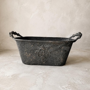 Op de afbeelding: Een ovale metalen plantenbak met een donkere, verweerde antracietkleurige afwerking en een verouderde textuur. De bak is voorzien van twee zijhandgrepen met een kralenontwerp en een verhoogd, sierlijk bloemenembleem op de voorzijde.