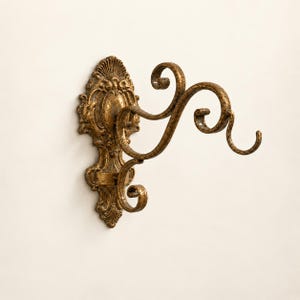 May include: A wall-mounted hook featuring an ornate, gold-toned metal backplate with intricate scrollwork and a matching curved arm that extends outward, ending in a decorative hook for hanging items, finished with a distressed, aged texture.