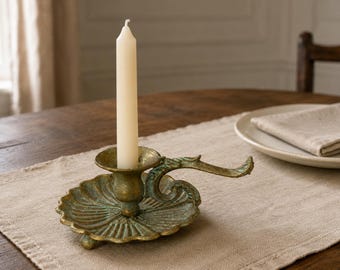 Puede incluir: Un candelabro de latón de estilo antiguo con una base festoneada en forma de hoja y un asa curva. El acabado metálico presenta una pátina moteada en verde y dorado. Una vela cónica de color blanco roto se mantiene erguida en el centro.