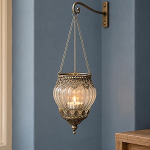 May include: A hanging candle holder featuring a clear, ribbed glass bowl encased in an ornate, antique brass-toned metal frame with filigree detailing. The bowl is suspended by three thin metal chains from a matching wall-mounted bracket arm.