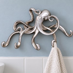 May include: A silver-toned octopus-shaped wall hook with multiple curved tentacles designed for hanging items. The metallic finish has subtle dark shading to highlight the textured suckers and the form of the octopus head and arms.