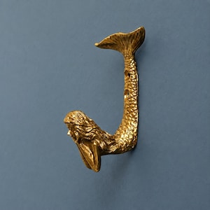 Könnte beinhalten: Ein goldfarbener Wandhaken in Form einer Meerjungfrau. Der Haken zeigt eine detaillierte Meerjungfrauenfigur mit langem Haar und einem strukturierten Schwanz, der sich nach oben biegt, um den Haken zu bilden. Zwei Schraublöcher sind sichtbar.